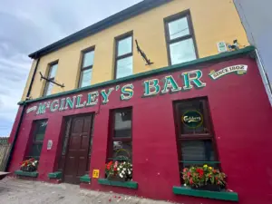 The front of McGinley’s Bar, established in 1802, with red and yellow walls, flower boxes under windows, and signs for Carlsberg and Jameson.