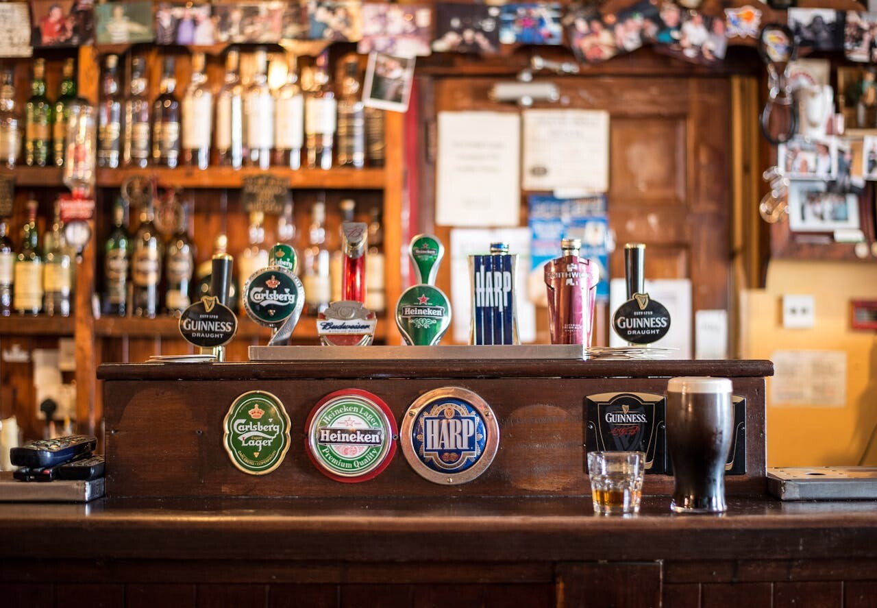 A classic Ireland bar counter features beer taps for Guinness, Carlsberg, Heineken, and Harp, surrounded by liquor bottles, photos, and memorabilia. A pint and a glass are pictured as well.