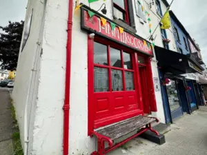 A red-painted storefront with a bench in front, colorful flags hanging above, and a sign that reads “S-NN INN LISC. COOB.” The building is on a sloped street with other shops nearby.