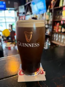 A Guinness pint glass with a creamy head sits on a coaster atop a dark bar counter in a cozy pub, with shelves of bottles and blurred TV screens visible in the background.