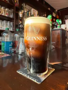 A Guinness pint glass with a creamy head sits on a bar counter, reflecting light. The Guinness logo is visible on the glass, and shelves with bottles and glasses are in the background, evoking a cozy pub atmosphere.