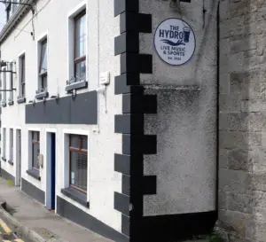 A building corner with a sign reading “The Hydro Live Music & Sports Est. 2023.” The building has white walls with black trim and multiple windows; a stone wall is on the right.