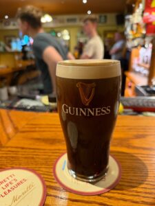 A Guinness pint glass with a thick, creamy head sits on a coaster atop a wooden pub bar, with two people blurred in the background and various bottles and bar items visible.