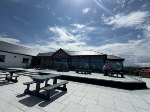 A sunny outdoor patio with dark wooden picnic tables, several people sitting and eating, and a modern building with large windows in the background under a partly cloudy sky—a classic scene outside a pub with Guinness pint glasses on the tables.