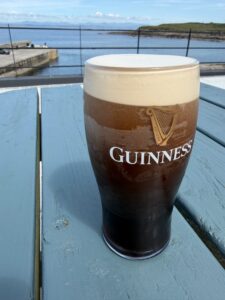 A Guinness pint glass with a creamy head sits on a blue wooden table at a pub, overlooking a calm harbor, railings, and distant land under a partly cloudy sky.