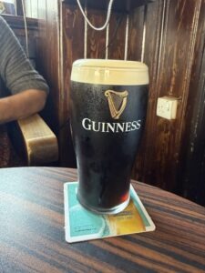 A pint of Guinness stout with a creamy head sits on a coaster atop a wooden table in a cozy pub setting. Part of a person's arm is visible resting on a wooden bench in the background.