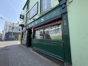 Street view of a green pub with dark green trim and signage reading "LEY'S." A large window displays a reflection of the street and an image of two Guinness pints, with buildings visible in the background.
