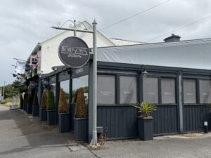 A dark blue restaurant called "Harrisons" with large windows, potted plants outside, and a hanging round sign. The text on the windows advertises al fresco dining and cocktails. The sky is cloudy.