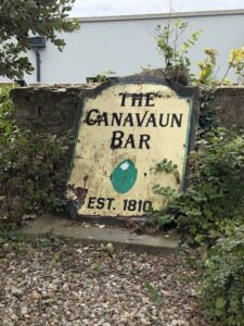 A weathered sign reading "The Canavaun Bar EST. 1810" rests against a stone wall, surrounded by overgrown plants and gravel. The sign is faded and damaged, with peeling paint and visible cracks.