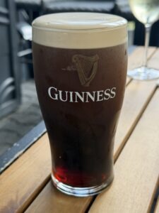 A pint glass of Guinness stout with a creamy head sits on a wooden table outdoors. The Guinness logo with a harp is visible on the glass. Another drink is blurred in the background.