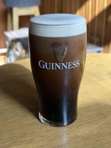 A pint glass of Guinness stout sits on a wooden table in sunlight, showcasing its dark body and creamy white head. The Guinness logo with a harp is printed on the glass.