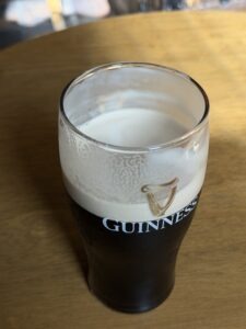 A pint glass of Guinness stout, mostly full with a creamy head, sits on a wooden table. The glass features the Guinness logo with a harp.