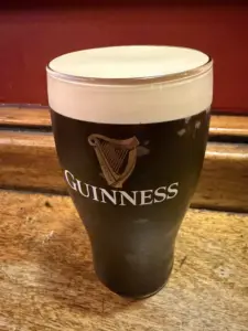 A pint glass filled with Guinness stout, topped with a creamy foam head, sits on a wooden surface with a red wall in the background. The Guinness logo and harp are visible on the glass.
