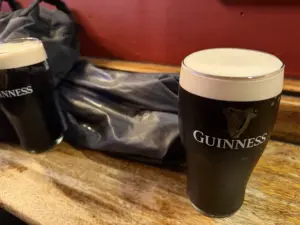 Two pints of Guinness stout on a wooden table, one in the foreground and one partially visible in the background, with a black jacket draped on the bench and a red wall behind.