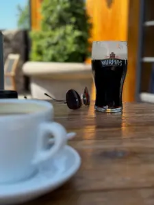 A glass of Murphy’s stout, a cup of coffee, and a pair of sunglasses rest on a wooden outdoor table in a sunlit setting with greenery in the background.