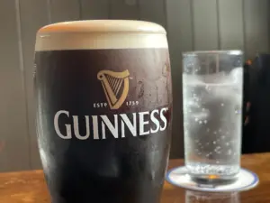 A Guinness pint glass with a creamy head sits on a wooden table next to a clear glass of sparkling water on a coaster, set against a dark, paneled pub background.