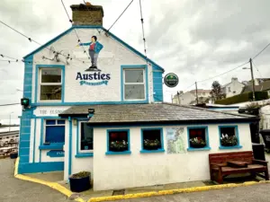 A seaside pub called Austies, with white walls, blue trim, and a mural of a sailor on the upper front. Benches sit outside alongside flower boxes under the windows, and a Guinness pint glass pub sign hangs nearby beneath the overcast sky.