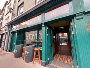 Green pub exterior with gold accents, two wooden stools, and large black barrels as tables on the sidewalk. Entrance door is open, revealing a tiled floor and some signage inside. The number 30 is above the doorway.