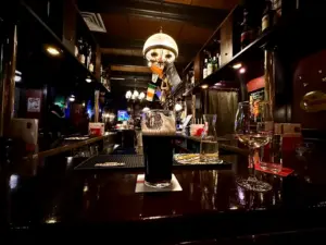 A dimly lit pub features a Guinness pint glass, a glass of white wine, and a carafe on the polished counter. Bottles line the shelves, and flags hang above the bar under warm hanging lights.