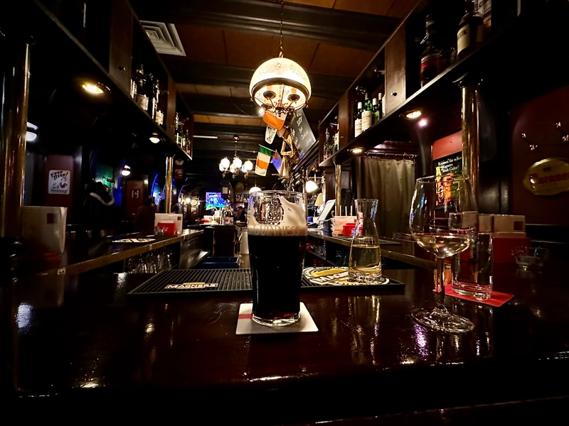 A dimly lit pub features a Guinness pint glass, a glass of white wine, and a carafe on the polished counter. Bottles line the shelves, and flags hang above the bar under warm hanging lights.