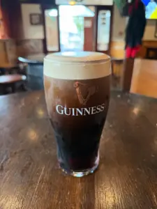 A Guinness pint glass with a creamy head rests on a wooden table inside a cozy pub, with the entrance and windows softly blurred in the background.