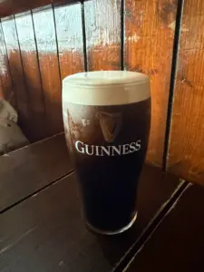 A Guinness pint glass sits on a dark wooden table in a cozy pub, featuring a thick creamy head and the iconic Guinness logo on the glass.