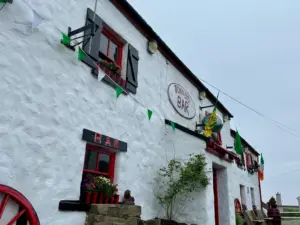 A white stone building with red-trimmed windows, green and white bunting, and a sign reading "Bonner’s Bar." Flower boxes, a small Irish flag, and a classic Guinness pint glass decorate the pub's exterior on a cloudy day.