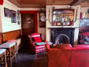 Cozy room with red leather armchairs, a fireplace, mirror, bookshelf, wooden stools, framed photos, a clock on the mantel, and a Guinness pint glass on the table, creating a warm, vintage pub atmosphere.