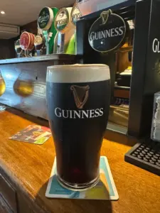 A Guinness pint glass sits on a coaster at a wooden pub bar, with taps—including Guinness and Carlsberg—visible in the background.