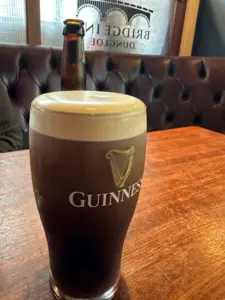 A Guinness pint glass with a creamy head sits on a wooden table in a pub, framed by tufted leather seating and a brown glass bottle. Sunlight pours through a window adorned with "Bridge" lettering in the background.