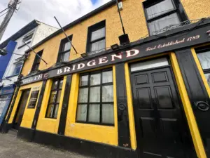 A yellow and black pub building with the sign "The Bridgend" and "First Established 1789" above the windows and doors. The angled view shows the front facade, some adjacent buildings, and a guinness pint glass on a nearby table.