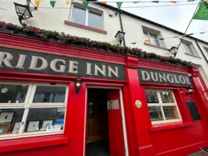 The exterior of a bright red pub called "Bridge Inn Dungloe," with flower boxes above the entrance, black trim, bunting overhead, and a Guinness pint glass displayed in the window. The door is open, with framed items visible through the windows.