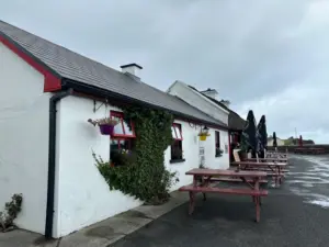 A white cottage-style building with red trim and a sloped roof features ivy, flowerpots, and wooden picnic tables with umbrellas outside on a cloudy day. The ground is wet, suggesting recent rain.