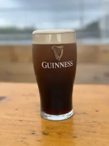 A pint glass filled with Guinness stout sits on a wooden table. The beer has a foamy, creamy head and the iconic Guinness logo is visible on the glass. The background is softly blurred.