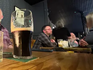 A nearly finished pint of dark beer on a wooden table in a dimly lit pub, with people sitting and talking in the background.