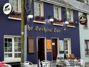 The exterior of The Beehive Bar, a purple building with flower boxes, outdoor seating, and signs for Carling and Guinness. Several lamps hang over the entrance, and a Guinness pint glass decorates the pub’s welcoming facade.