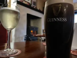 A glass of white wine and a Guinness pint glass rest on a wooden table in a cozy pub, with a lit fireplace and framed pictures adding to the inviting atmosphere.