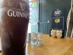 A Guinness pint glass on a wooden table in a pub, with a glass candle holder, ceramic bird salt and pepper shakers, and a "Boat Inn" menu visible in the background.