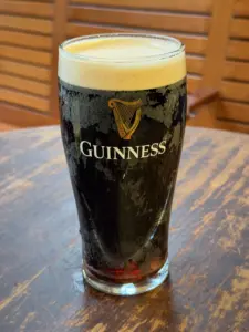 A pint glass of Guinness stout with a creamy head sits on a rustic wooden table, with a blurred wooden background.