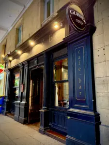 A dark blue cafe bar entrance with gold lettering, a round Guinness sign above, and large windows. The building has ornate details and appears to be on a city street with a covered walkway.