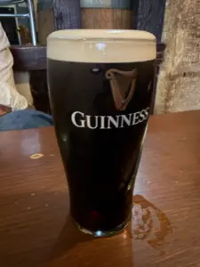 A Guinness pint glass with a creamy head sits on a wooden table in a cozy pub setting. The Guinness logo and harp emblem are visible on the glass.