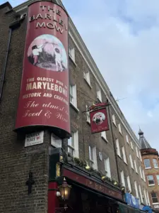 A brick building with a red sign reading "The Barley Mow, The Oldest Pub in Marylebone," featuring an illustration of a dog. The pub is on Enrick Place, with festive decorations above the entrance.
