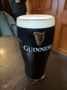 A pint of Guinness stout with a creamy head sits on a dark wooden table in a pub, with a wooden paneled wall and a partially visible person in the background.
