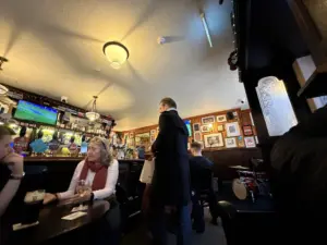 A lively pub scene with people sitting at tables and chatting. The bar is visible with drinks and a TV showing a soccer game. Framed pictures line the walls, and a small drum set is set up in the corner.