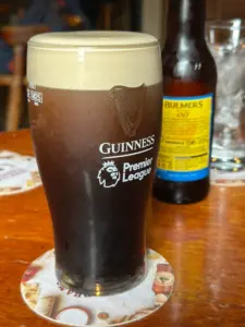 A Guinness pint glass with a creamy head sits on a coaster on a wooden table in a pub, with a bottle of Bulmers cider and an empty glass with ice in the background.