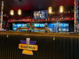 A bar counter with a "Le Brasstorium" sign, guinness pint glass, beer taps, a colorful spinning wheel, and shelves stocked with bottles and cans behind. A menu board is displayed above the pub bar. Warm hanging lights illuminate the scene.