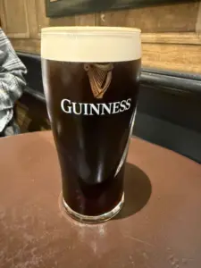 A pint glass of Guinness stout with a creamy head sits on a brown table, with wooden paneling and part of a person's arm visible in the background.