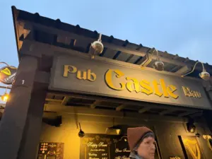 A person in a brown hat stands in front of a pub with a large sign reading "Pub Castle Point" in gold letters. The exterior is dark with hanging lamps and a Guinness sign visible. It appears to be early evening.
