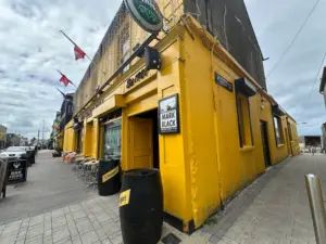 A bright yellow pub with outdoor seating and barrels on the sidewalk. Signs and a poster that says "Mark Black" are visible, along with a Guinness pint glass in the window. Flags hang above the street under a cloudy sky.