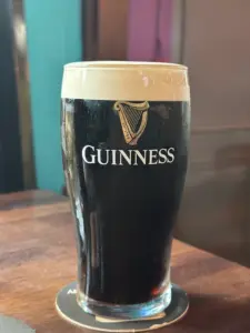 A Guinness pint glass with a creamy head sits on a wooden table at the pub, light shining through the dark stout. The iconic Guinness logo and harp symbol are visible on the glass.
